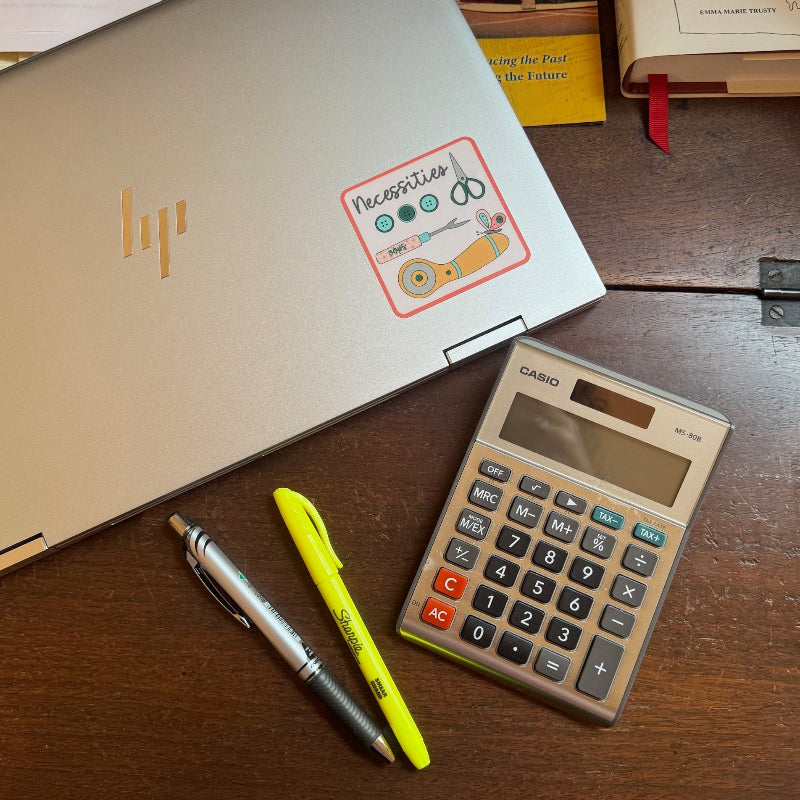 Silver laptop on desk with a sticker featuring sewing notions and the word Necessities.