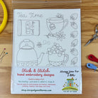 A clear plastic sleeve containing a white fabric design sheet with black line drawings of tea-themed motifs, including a teapot, teacup, flowers, tea bag, and strawberries, plus printed product text. The sheet is photographed on a wooden surface with sewing supplies like pins, scissors, thread, and ribbon around it.