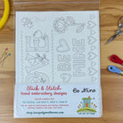 A packaged stick and stitch hand embroidery design kit featuring valentine themed 'Be Mine' designs, displayed on a wooden tabletop with embroidery floss, pins, and scissors nearby.
