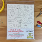 A clear plastic sleeve contains a sheet of hand embroidery designs featuring black line drawings of dogs, hearts, bones, and paw prints with the text 'Home is where the Dog is.' The bottom of the sheet has product information and branding for 'The Art of Home' and 'Stik & Stitch hand embroidery designs.