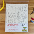 A clear plastic bag containing a hand embroidery design kit sheet labeled 'Stick & Stitch hand embroidery designs' and 'Needle & Thread' by The Art of Home, placed on a wooden surface with sewing tools around it.