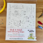 A packaged sheet of embroidery designs labeled 'Oh, Holy Night' and 'Stick & Stitch hand embroidery designs,' shown on a wooden surface with sewing tools like a yellow hoop, pins, scissors, and thread around it.