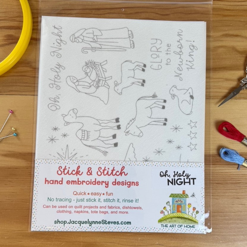 A packaged sheet of embroidery designs labeled 'Oh, Holy Night' and 'Stick & Stitch hand embroidery designs,' shown on a wooden surface with sewing tools like a yellow hoop, pins, scissors, and thread around it.