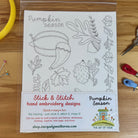A packaged 'Pumpkin Season' hand embroidery design kit by The Art of Home, featuring black and white line art of pumpkins, acorns, and autumn leaves on a white fabric backing, displayed on a wooden table with sewing tools.