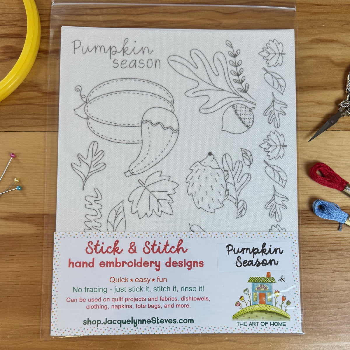 A packaged 'Pumpkin Season' hand embroidery design kit by The Art of Home, featuring black and white line art of pumpkins, acorns, and autumn leaves on a white fabric backing, displayed on a wooden table with sewing tools.