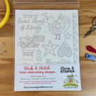 A sheet of hand embroidery designs in a clear plastic sleeve on a wooden table, with embroidery floss, scissors, and pins around it. The sheet reads 'Sweet Land of Liberty' and shows a bird, stars, flowers, and hearts in outline embroidery patterns.