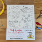 A packaged sheet of hand embroidery designs featuring winter illustrations and the text 'COZY WINTER WISHES,' shown on a wooden surface with embroidery floss, pins, and scissors around it. The package label reads 'Stick & Stitch hand embroidery designs' and 'Warm Winter Wishes,' with 'The Art of the House' branding.