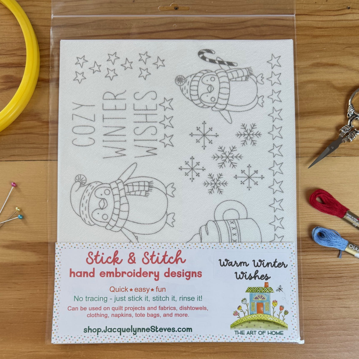 A packaged sheet of hand embroidery designs featuring winter illustrations and the text 'COZY WINTER WISHES,' shown on a wooden surface with embroidery floss, pins, and scissors around it. The package label reads 'Stick & Stitch hand embroidery designs' and 'Warm Winter Wishes,' with 'The Art of the House' branding.