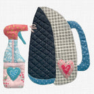 A handmade quilted applique iron and spray bottle set on a white grid background. The iron has a navy blue polka dot section, a gray and white gingham section, and a blue handle with a pink heart applique.