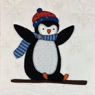 A quilted applique design of a penguin wearing a red and blue knit hat and a blue striped scarf, sitting on a brown sled against an off-white fabric background with embroidered snowflakes.