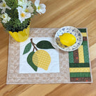 A square quilted placemat with a lemon design, styled on a light wood tabletop next to a potted white flower and a small bowl containing a lemon.