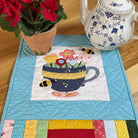 A quilted fabric table runner with a colorful patchwork border and a central floral teacup design, styled on a wooden table next to a potted red flower and a blue-and-white teapot.