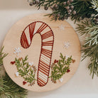 Wooden round board with embroidered candy cane design surrounded by greenery