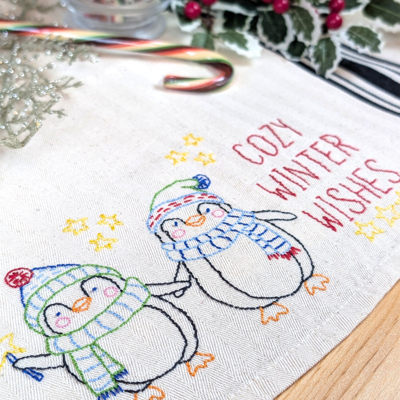 A close-up of a white woven table runner with embroidered penguins in winter hats and scarves, yellow stars, and the words 'COZY WINTER WISHES' in red. The runner is styled on a wooden surface with holiday decorations including a candy cane, holly, and a glass of striped drink.