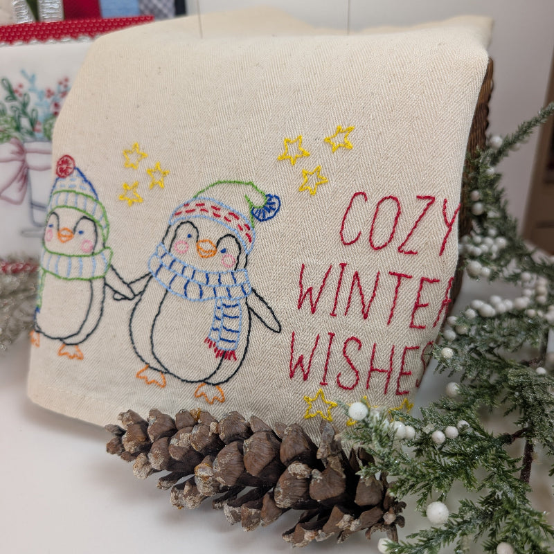 A square cream linen fabric napkin or small tablecloth with embroidered penguins in winter hats and scarves, yellow stars, and the words 'COZY WINTER WISHES' in red. The fabric is styled with pinecones and evergreen branches on a white surface.