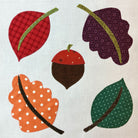 A square with applique design featuring five autumn-themed applique shapes: two leaves, an acorn, and two additional leaves, each with different patterns like plaid, polka dots, and floral prints on a light beige linen background.