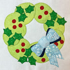 A handmade fabric applique wreath featuring light green and dark green holly leaves with red berries, finished with a light blue patterned bow on a white fabric background.