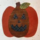 A handmade fabric applique of a jack-o'-lantern pumpkin. The body features a brown and black houndstooth pattern for the face, with solid rust-orange felt sides, a matching rust-orange felt stem, and a green houndstooth leaf on top, all set against a cream fabric background with small gold star prints.