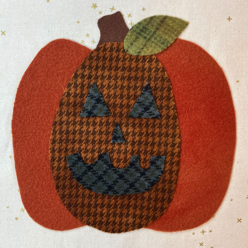 A handmade fabric applique of a jack-o'-lantern pumpkin. The body features a brown and black houndstooth pattern for the face, with solid rust-orange felt sides, a matching rust-orange felt stem, and a green houndstooth leaf on top, all set against a cream fabric background with small gold star prints.