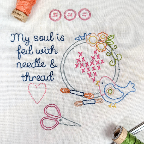 A close-up of a white fabric surface with embroidered text reading 'My soul is fed with needle & thread' and a decorative motif including a blue bird, pink flowers, scissors, buttons, and thread spools. No person or real environment is present, so this is Product/Packaging rather than Lifestyle.