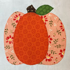 A handmade fabric applique pumpkin with a solid orange-red center, a peach floral side, and a green leaf on a light beige linen background.