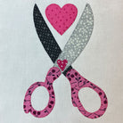 A close-up of a fabric applique or printed design featuring crossed scissors with a pink heart above them, on a light beige fabric background.