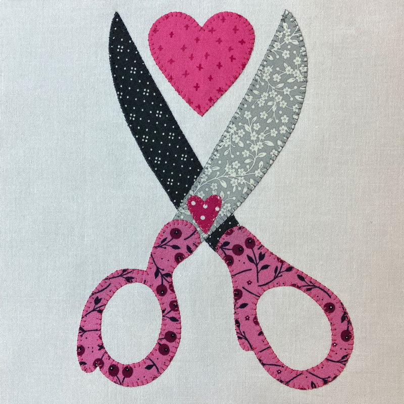 A close-up of a fabric applique or printed design featuring crossed scissors with a pink heart above them, on a light beige fabric background.